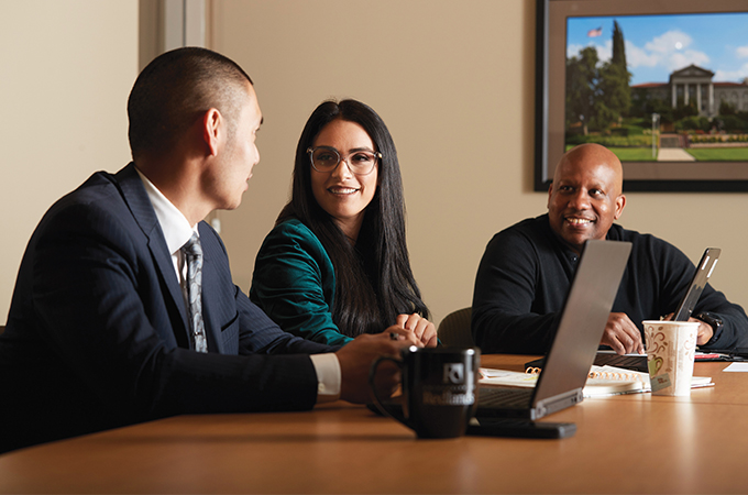 A woman and a man sitting at a table with laptops talk to each other while another man looks on.